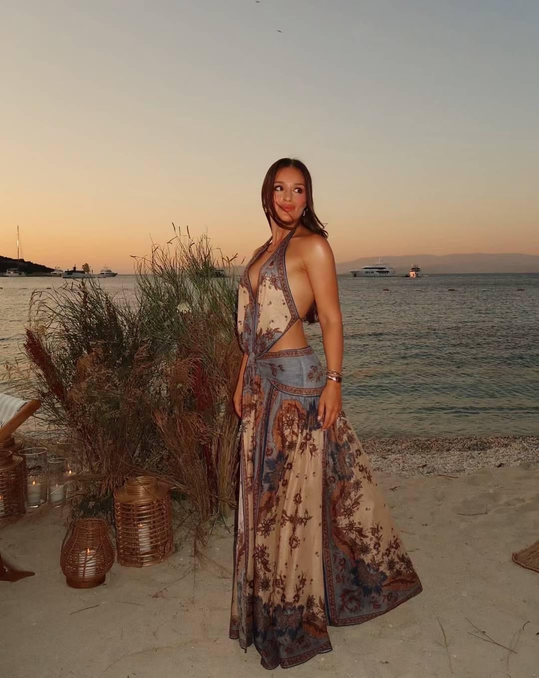 Vestido largo sin mangas, con cuello halter y nudo delantero a la altura de la cintura. Presenta un diseño fluido con abertura frontal y estampado tipo bohemio en tonos beige, marrón y azul grisáceo